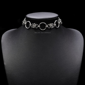 Puede incluir: Un collar gargantilla plateado con una serie de anillos conectados y elementos decorativos. El collar se exhibe en un maniquí negro. El texto "ARCANE ACCESSORIES" es visible.