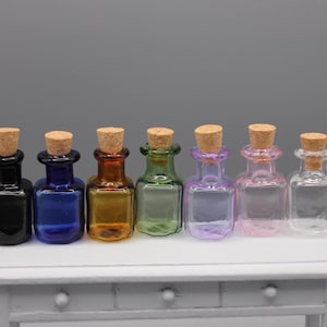 May include: Seven miniature glass bottles with cork stoppers, each containing a different coloured liquid. The bottles are in shades of black, blue, amber, green, purple, pink, and clear. They are displayed on a small white table.