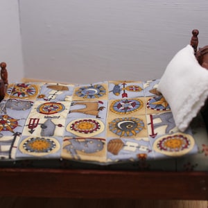 May include: A miniature wooden bed frame with a dark brown finish, featuring a small white pillow and a quilted blanket. The blanket displays a repeating grid pattern of colourful illustrations, including suns, moons, compasses, and various animal figures.
