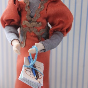 May include: A doll wearing a red dress with a brown lace trim and gray sleeves holds a small blue notebook with two pencils tied with blue string. The notebook has the words "Exam Book" printed on the cover.