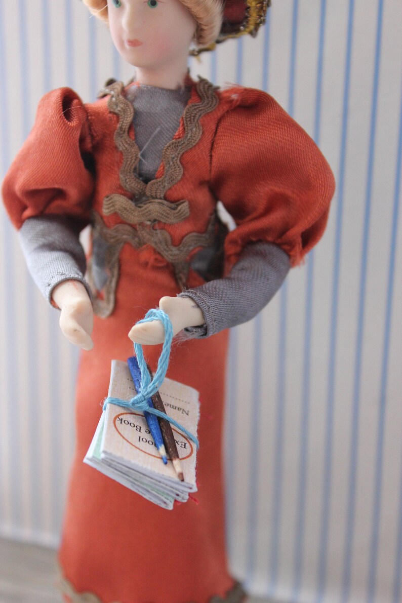 May include: A doll wearing a red dress with a brown lace trim and gray sleeves holds a small blue notebook with two pencils tied with blue string. The notebook has the words "Exam Book" printed on the cover.