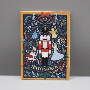 May include: Rectangular card with a navy blue background and a gold border. The card features a festive illustration of a nutcracker, angel, and ballerina, with the text "The Nutcracker" at the bottom. The design includes Christmas-themed elements.