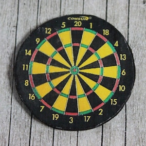 May include: A black and yellow dartboard with red and green accents. The numbers 1 through 20 are printed around the board, along with the words "COMSUN" at the top.