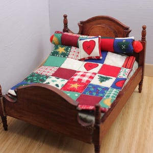 May include: A miniature wooden bed frame with a dark brown finish, featuring a patchwork quilt and a matching bolster pillow. The bedding displays a festive pattern of red, green, and white squares with holiday motifs, including a small heart pillow.