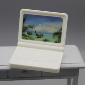 May include: A small white laptop showing a tropical beach scene with boats. The laptop has a keyboard and is open, placed on a white table. The screen displays a blue sky and turquoise water.