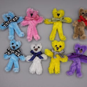 May include: Eight colourful pipe cleaner teddy bears, each with a bow or ribbon. The bears are in shades of blue, pink, yellow, brown, white, and purple. They have black bead eyes and are set against a grey backdrop.