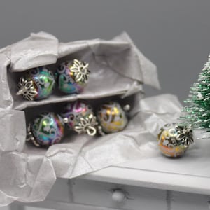 May include: Miniature Christmas baubles in a box. The baubles are round, with iridescent colours and silver-coloured floral caps. The box is lined with white tissue paper, and a small Christmas tree is in the background.
