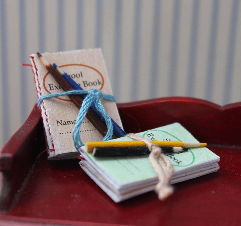 May include: Two miniature school books with pencils on a red surface. The larger book is red and has the text "School Exercise Book" and "Name" on the cover. The smaller book is green and has the text "School Exercise Book" on the cover.