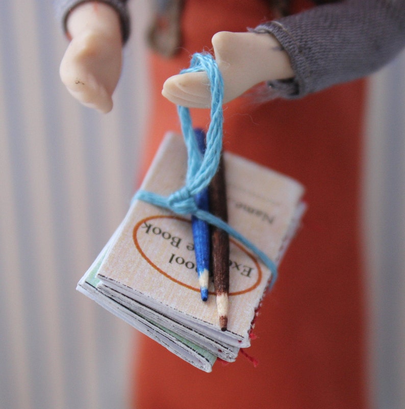 May include: A stack of small, cream-colored books tied together with a blue string. The books have the text "Excel" and "Cool Book" printed on the covers. Two wooden pencils are tucked into the string.