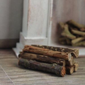 May include: A miniature fireplace scene featuring a stack of small, weathered wooden logs. The logs are brown and tan, with visible bark and texture. The background includes a white fireplace mantel and a pile of similar logs.