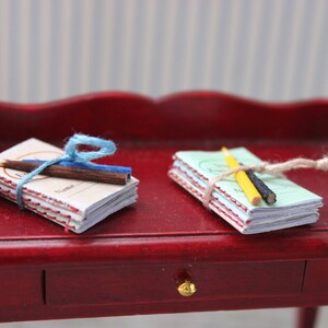 May include: Two stacks of miniature notebooks tied with twine and decorated with pencils. The notebooks are on a red wooden surface.