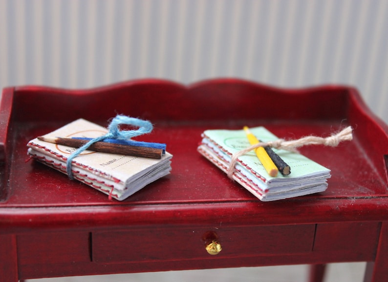 May include: Two stacks of miniature notebooks tied with twine and decorated with pencils. The notebooks are on a red wooden surface.