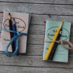 May include: Two miniature school exercise books with pencils tied around them. The books are green and white with red and white trim. The pencils are yellow and blue.