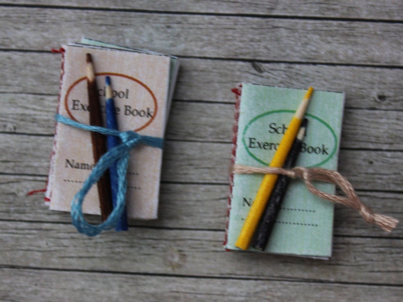May include: Two miniature school exercise books with pencils tied around them. The books are green and white with red and white trim. The pencils are yellow and blue.