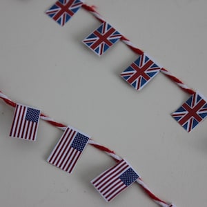 May include: Two strings of miniature flags, one with the Union Jack and the other with the American flag. The flags are attached to a red and white twisted cord, set against a white background.