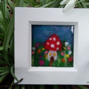 Op de afbeelding: Een ingelijst naaldviltwerk dat een rood en wit paddenstoelenhuis met een witte deur en twee gele ramen toont. Het paddenstoelenhuis is omgeven door groen gras en kleurrijke bloemen.