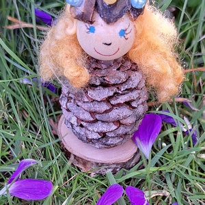 May include: A whimsical fairy doll crafted from a pine cone, featuring a painted face, blonde yarn hair, and blue and gold embellishments. The doll is nestled amongst green grass and purple flower petals.