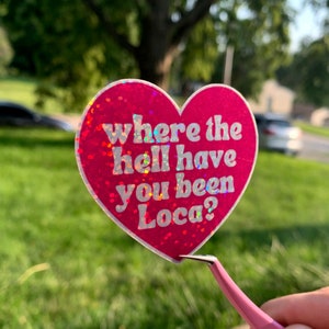 May include: A heart-shaped sticker with a pink glitter background and the text "where the hell have you been Loca?" in white. The sticker is held by pink tweezers against a green grassy background.