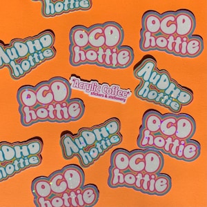May include: A collection of stickers on an orange surface. The stickers display the phrases "OCD hottie" and "AuDHD hottie" in a bubble font with rainbow and glitter details. A small sticker reads "Acrylic Coffee stickers & stationery."