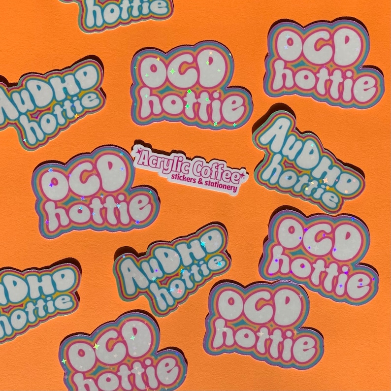 May include: A collection of stickers on an orange surface. The stickers display the phrases "OCD hottie" and "AuDHD hottie" in a bubble font with rainbow and glitter details. A small sticker reads "Acrylic Coffee stickers & stationery."