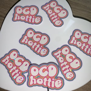 May include: Several stickers with the text "OCD hottie" in a bubble letter style. The stickers feature a white background with pink, orange, and blue outlines. Some stickers have a holographic, glittery finish.