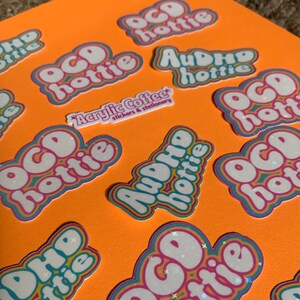 May include: A collection of stickers on an orange surface. The stickers display the phrases "OCD hottie" and "AuDHD hottie" in a bubble font with a rainbow border. A smaller sticker reads "Acrylic Coffee stickers & stationery."