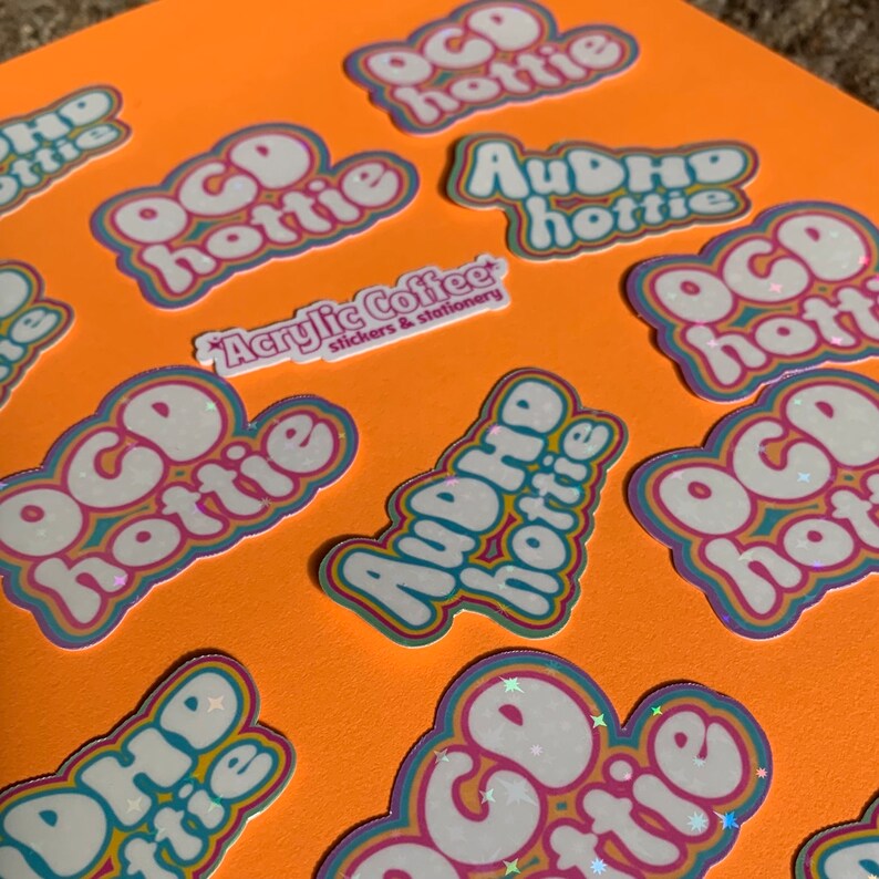 May include: A collection of stickers on an orange surface. The stickers display the phrases "OCD hottie" and "AuDHD hottie" in a bubble font with a rainbow border. A smaller sticker reads "Acrylic Coffee stickers & stationery."