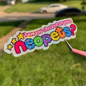 May include: A holographic sticker with the text "have you checked on your neopets?" in colorful bubble letters. The sticker has a wavy border and star accents. A pink tweezer is holding the sticker.