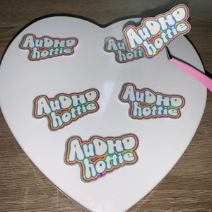 May include: A selection of stickers featuring the text "AuDHD hottie" in a bubble font, with rainbow-coloured outlines and a holographic finish. The stickers are displayed on a white heart-shaped surface.