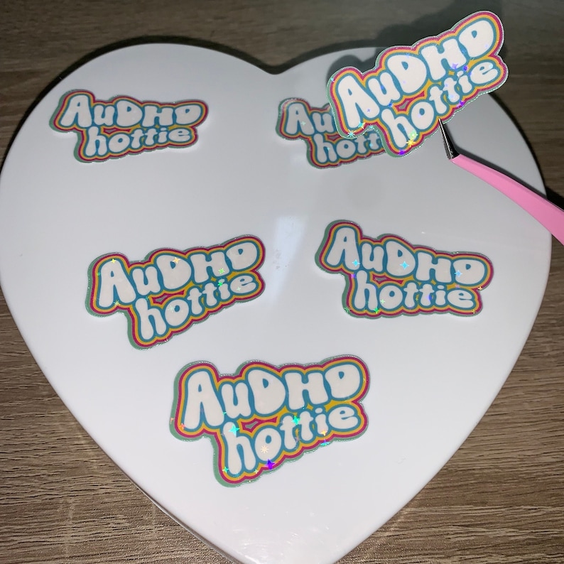 May include: A selection of stickers featuring the text "AuDHD hottie" in a bubble font, with rainbow-coloured outlines and a holographic finish. The stickers are displayed on a white heart-shaped surface.