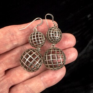 May include: Silver-toned drop earrings featuring a cage-like design with two spheres. The spheres have a grid pattern and a reddish-brown interior. The earrings are held in a hand against a black background.