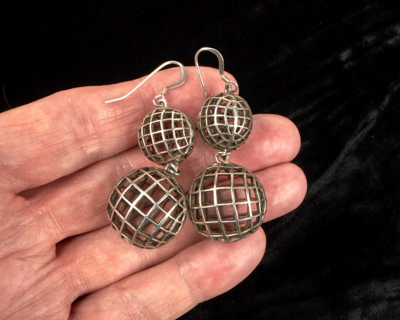 May include: Silver-toned drop earrings featuring a cage-like design with two spheres. The spheres have a grid pattern and a reddish-brown interior. The earrings are held in a hand against a black background.