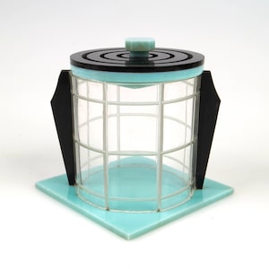 Può includere: Un barattolo di vetro in stile Art Déco con un coperchio. Il barattolo ha un design geometrico ed è realizzato in vetro trasparente con un bordo nero e blu.