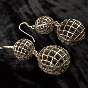 May include: A pair of silver-toned drop earrings. Each earring features two openwork spheres connected by a small chain. The spheres have a grid-like pattern, creating a cage-like design. The earrings are displayed on a black surface.