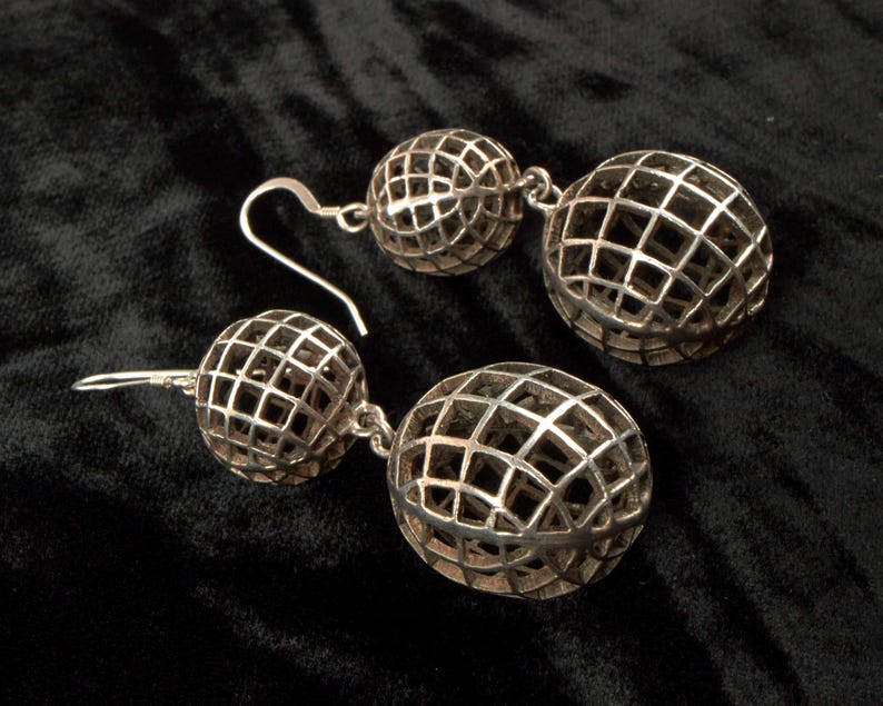 May include: A pair of silver-toned drop earrings. Each earring features two openwork spheres connected by a small chain. The spheres have a grid-like pattern, creating a cage-like design. The earrings are displayed on a black surface.