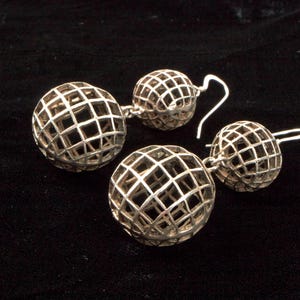 May include: Two pairs of silver-toned, openwork sphere earrings. Each earring features a grid-like design, creating a cage effect around the sphere. The earrings are displayed on a black surface, highlighting their metallic sheen.