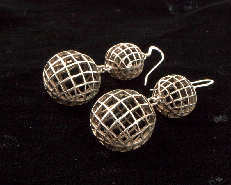 May include: Two pairs of silver-toned, openwork sphere earrings. Each earring features a grid-like design, creating a cage effect around the sphere. The earrings are displayed on a black surface, highlighting their metallic sheen.