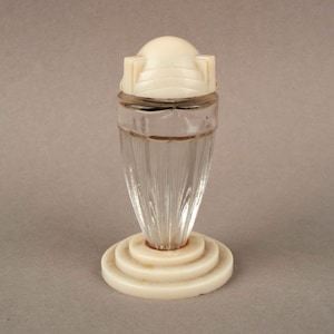 Può includere: Un'antica boccetta di profumo con un corpo in vetro trasparente scanalato e un tappo a cupola color crema. La bottiglia poggia su una base circolare a più livelli. Una sottile fascia dorata accentua la parte superiore del vetro. Il design ricorda l'epoca Art Déco.