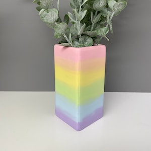 May include: A pastel rainbow coloured ceramic vase with a square shape. The vase is filled with a leafy green plant.