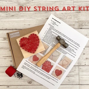 May include: A DIY string art kit featuring a red heart design. The kit includes a wooden base, nails, string, and a hammer. Instructions are printed on a sheet of paper.
