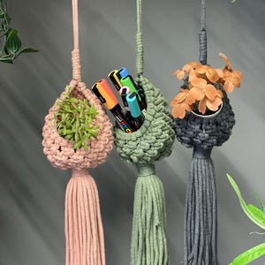 May include: Three hanging macrame plant holders in pink, green, and grey. The pink holder contains a small green plant, the green holder holds colourful markers, and the grey holder has orange flowers. Each has a tassel.
