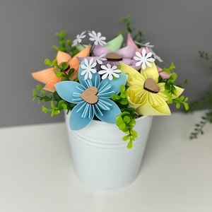 May include: A white metal bucket filled with colourful origami flowers. The flowers are in shades of blue, yellow, orange, and purple, with small white daisy accents and wooden heart centres. Greenery adds a natural touch to the arrangement.