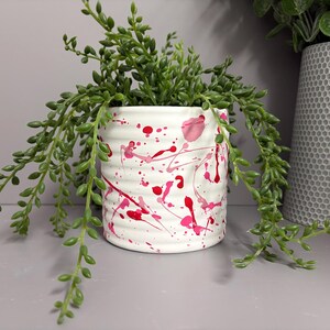 May include: A white ceramic plant pot with a textured finish, decorated with pink and red paint splashes. It contains a trailing green succulent plant. A grey vase with a honeycomb pattern is in the background.
