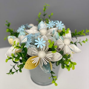 May include: A bouquet of white paper flowers with blue centres, made from maps, in a grey metal bucket. The flowers are surrounded by green leaves.