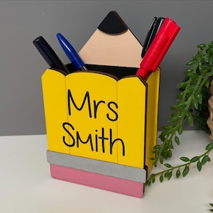 May include: A yellow pencil-shaped desk organiser with a pink eraser and the name "Mrs. Smith" written on the front. The organiser is holding three pens, one red, one blue, and one black.