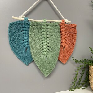 May include: A wall hanging made of macrame with three leaf shapes in teal, green, and orange colours. The leaves are connected by a wooden stick and hang from a white cord.