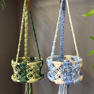 May include: Two macrame plant hangers featuring wooden rings at the top and base. One hanger is crafted from dark and light green cord, while the other uses blue and white cord. Both designs incorporate intricate knotted patterns and long tassels.