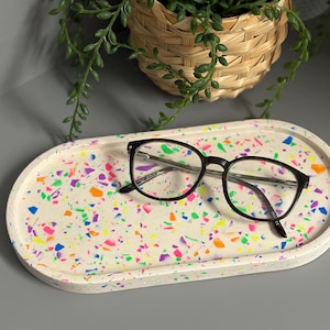 May include: An oval-shaped tray with a white base featuring a multi-coloured terrazzo pattern of neon pink, orange, blue, and green flecks. A pair of dark-rimmed spectacles with thin temples rests on the surface of the tray.