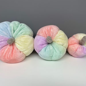 May include: Three plush pumpkins in various sizes, each with a different pastel colour scheme. The pumpkins are made of soft, fuzzy fabric and have a slightly textured surface.