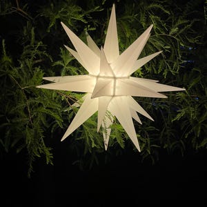 May include: A large, illuminated white star-shaped decoration with multiple points, hanging against a backdrop of dark green foliage. The star is lit from within, creating a soft glow. The overall theme is festive and decorative.